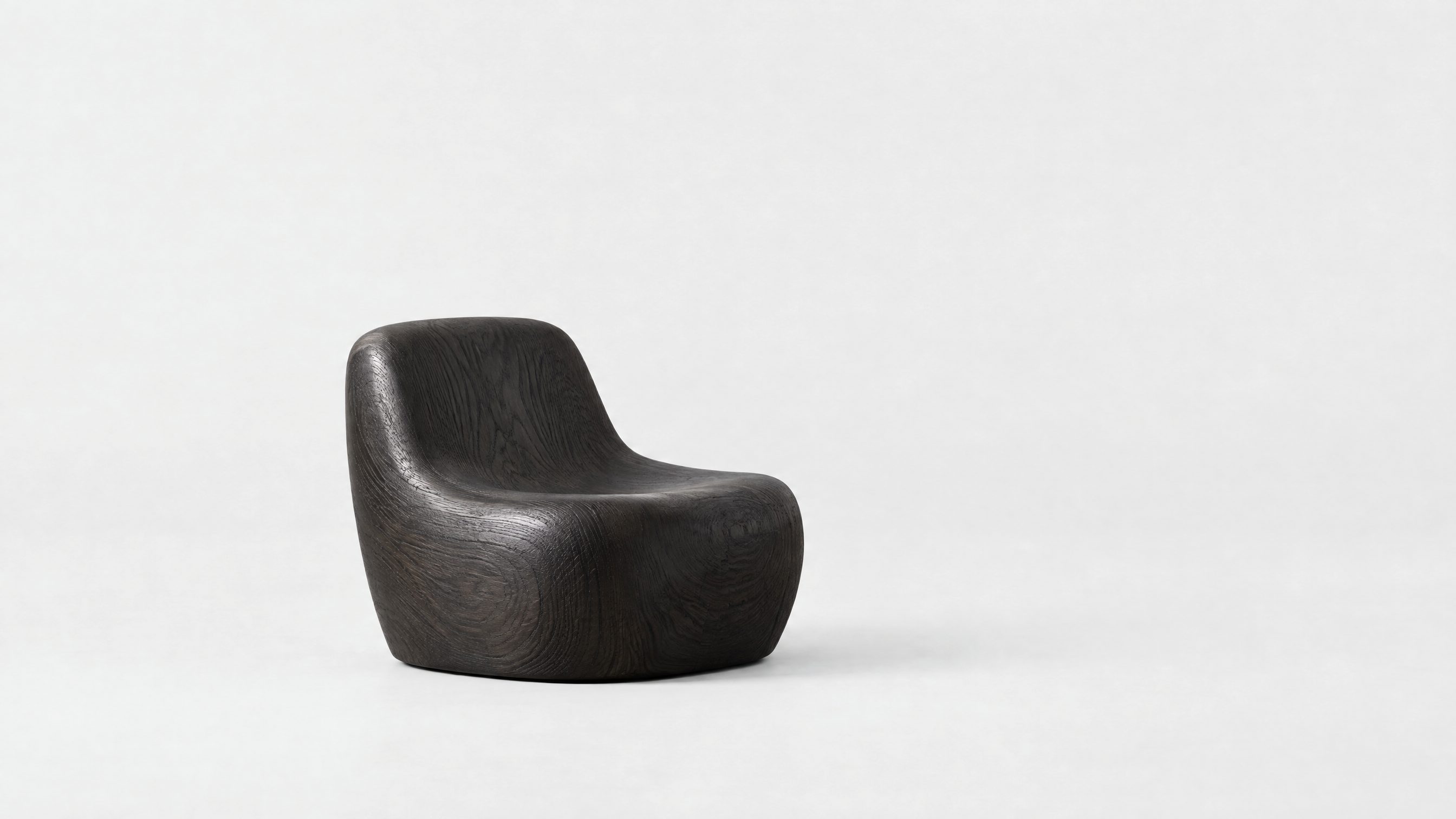 A single low armchair carved from black-stained oak, small in a wide, empty grey studio